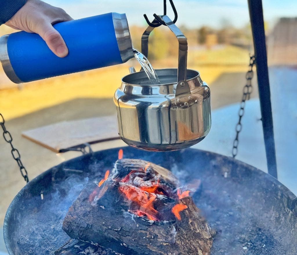A person pours water from a blue thermos into a stainless steel kettle hanging over a fire pit with burning logs. The scene is outdoors, with blurred surroundings indicating an open space or backyard.