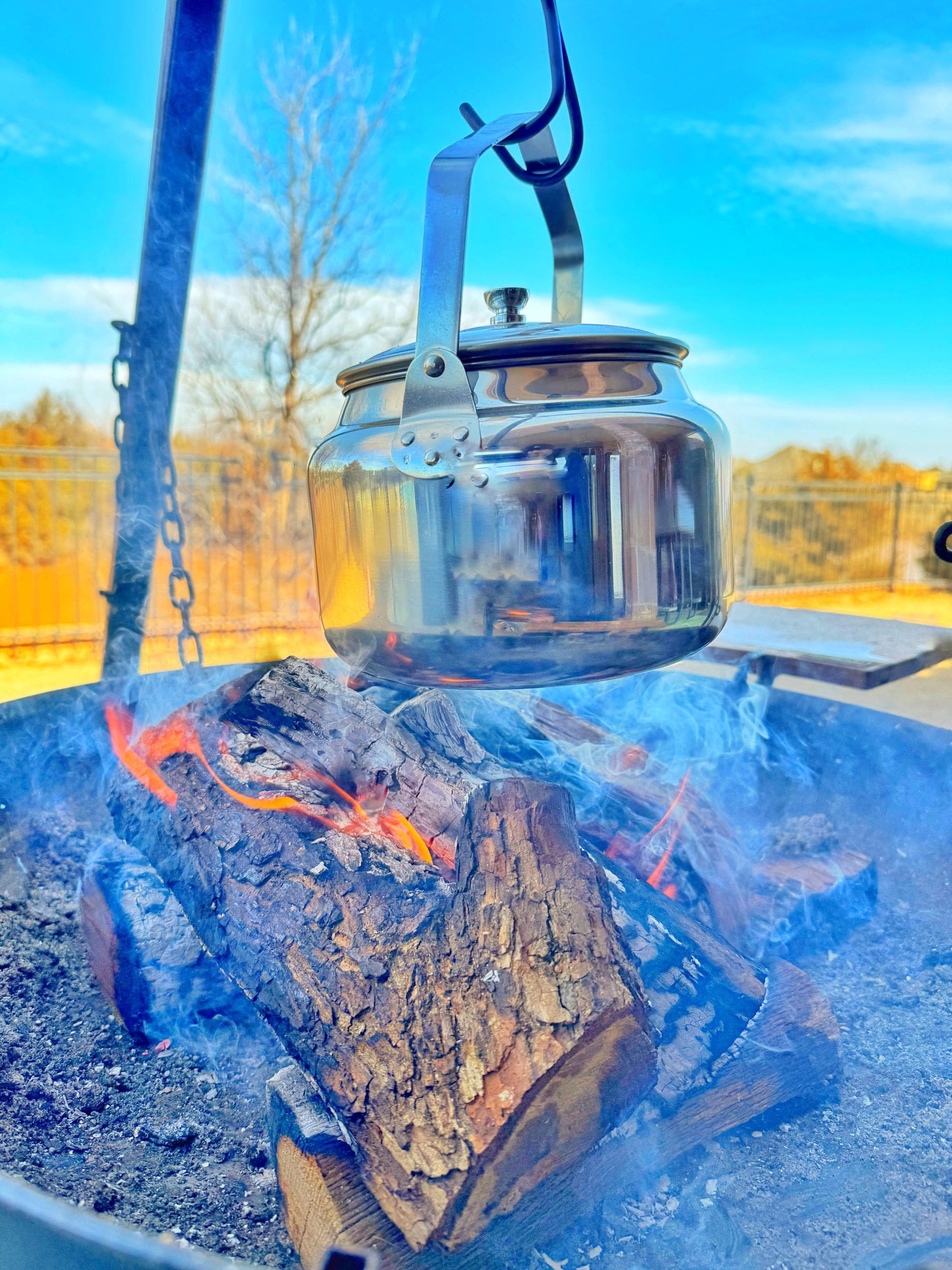 A metal pot hangs over a campfire on a tripod stand. The firewood is burning with visible orange flames and smoke. The background shows a clear blue sky and bare trees, suggesting an outdoor setting.