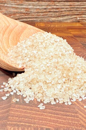 A wooden spoon lies on a wooden surface, spilling a heap of coarse sea salt crystals. The background is a textured wood pattern, enhancing the rustic appearance.