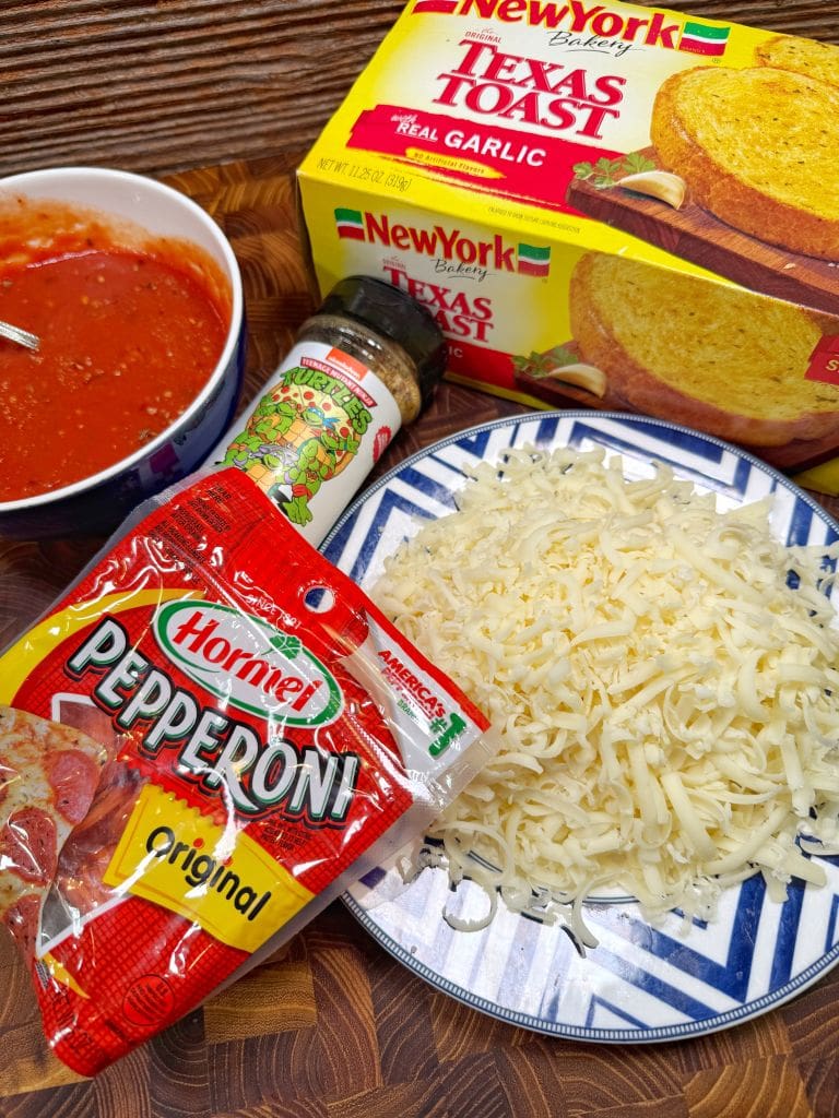 Ingredients for pizza bread on a wooden surface: a bowl of tomato sauce with a spoon, a package of New York Bakery Texas Toast garlic bread, a bottle of Italian seasoning, Hormel pepperoni pack, and shredded cheese on a blue plate. Getting ready to make garlic bread pizza