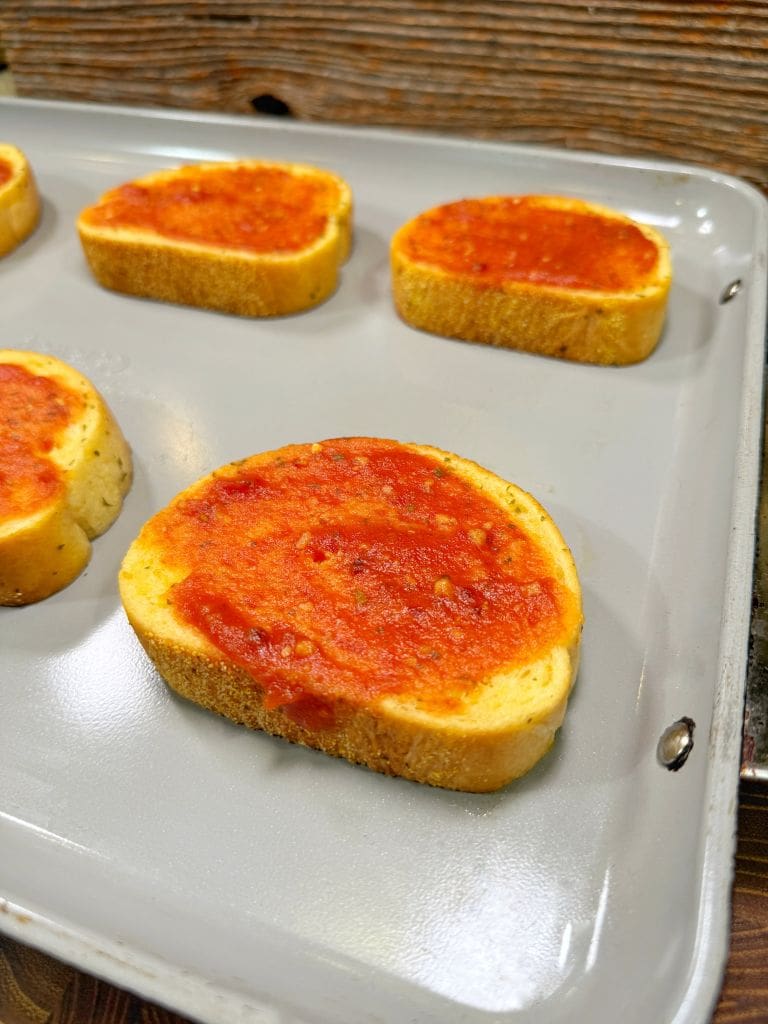 Slices of bread topped with tomato sauce on a baking tray. The tray is placed on a kitchen countertop.