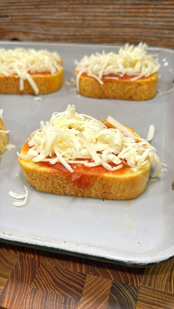 Slices of bread on a baking tray topped with tomato sauce and shredded cheese, ready to be baked. The tray is placed on a wooden surface.