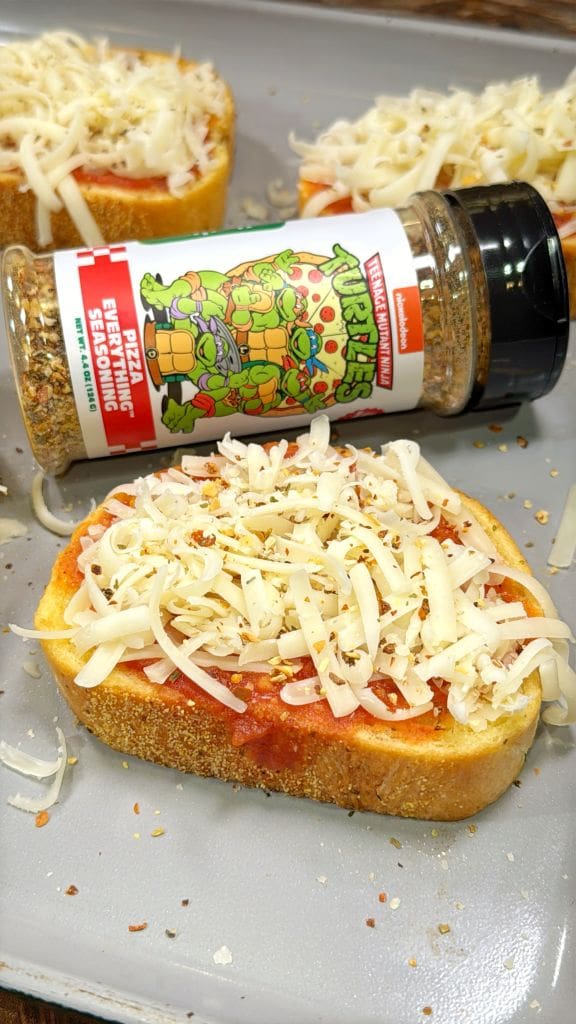 Slices of bread topped with tomato sauce and shredded cheese on a baking sheet. A bottle of Pizza Everything Seasoning, featuring a cartoon design and text in vibrant colors, is tipped over, with some seasoning sprinkled on the bread.
