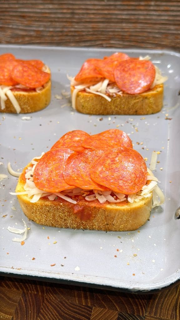 Slices of bread topped with tomato sauce, shredded cheese, and pepperoni are arranged on a baking sheet, ready to be baked.