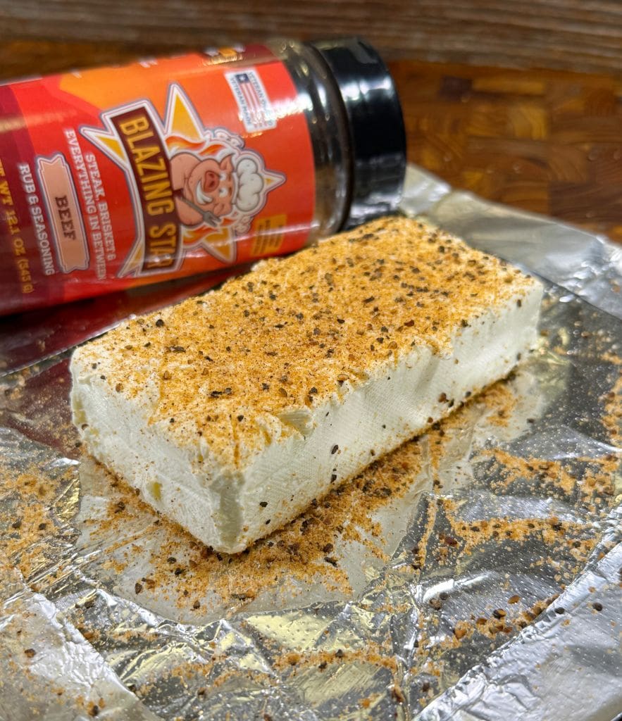 Block of cream cheese dusted with spicy seasoning on foil. A container of Blazing Star rub is tipped over in the background.
