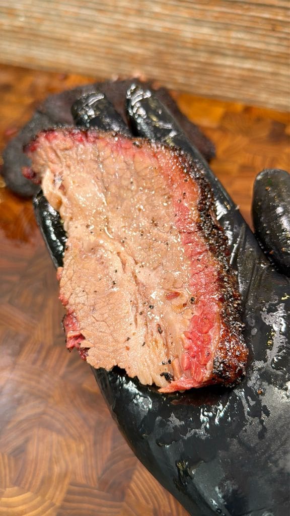 A gloved hand holds a slice of smoked brisket. The meat has a dark, seasoned crust and a tender, juicy interior with a red smoke ring. The wooden surface beneath the hand adds a rustic touch.