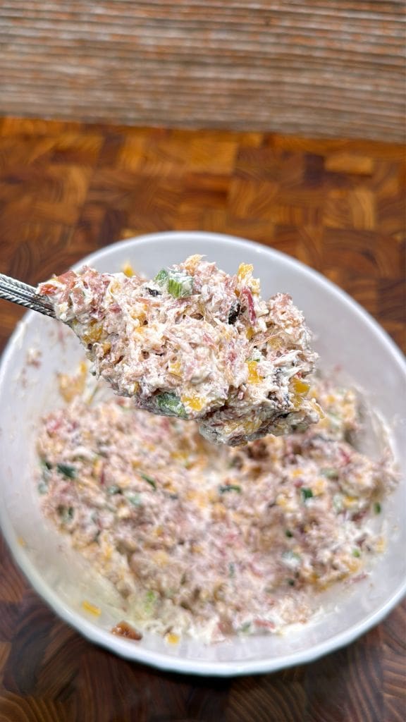 A spoon holds a portion of a creamy mixture with visible chopped vegetables and meat, possibly tuna or chicken salad, over a bowl filled with the same food. The background is a wooden surface with a textured pattern.