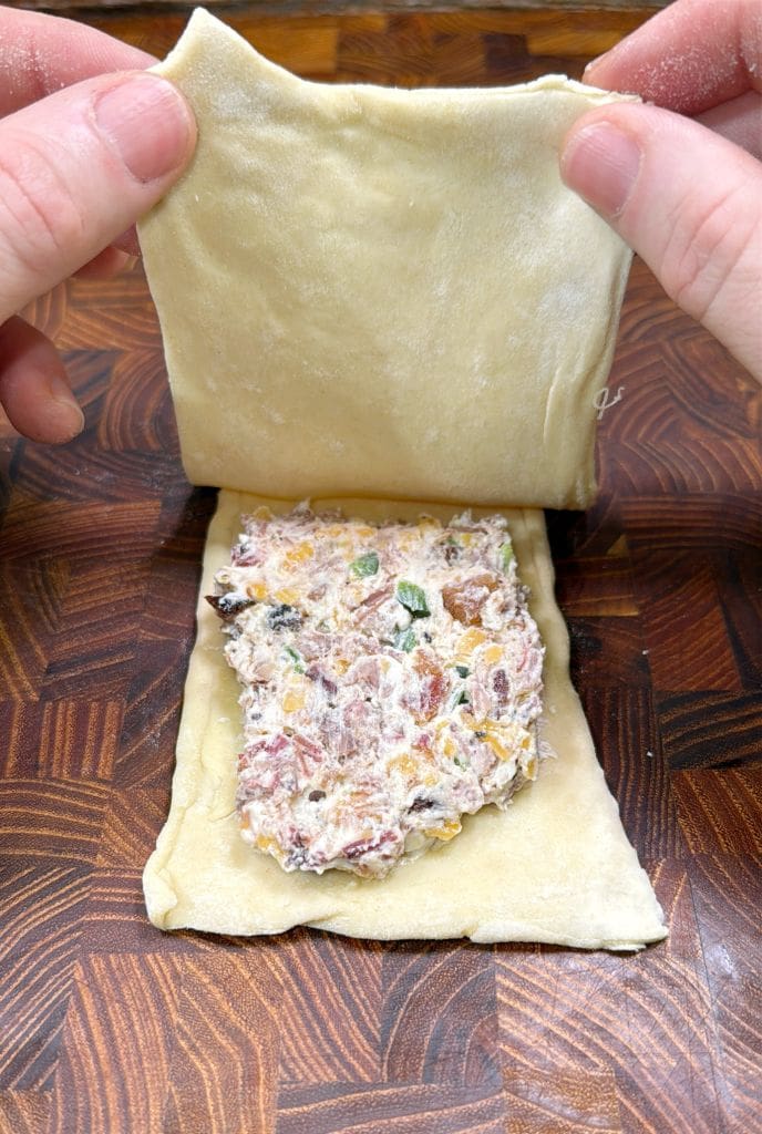 Hands are folding a sheet of dough over a filling mixture that includes visible green, red, and yellow ingredients. The preparation is on a wooden surface with a geometric pattern - holding homemade hot pockets