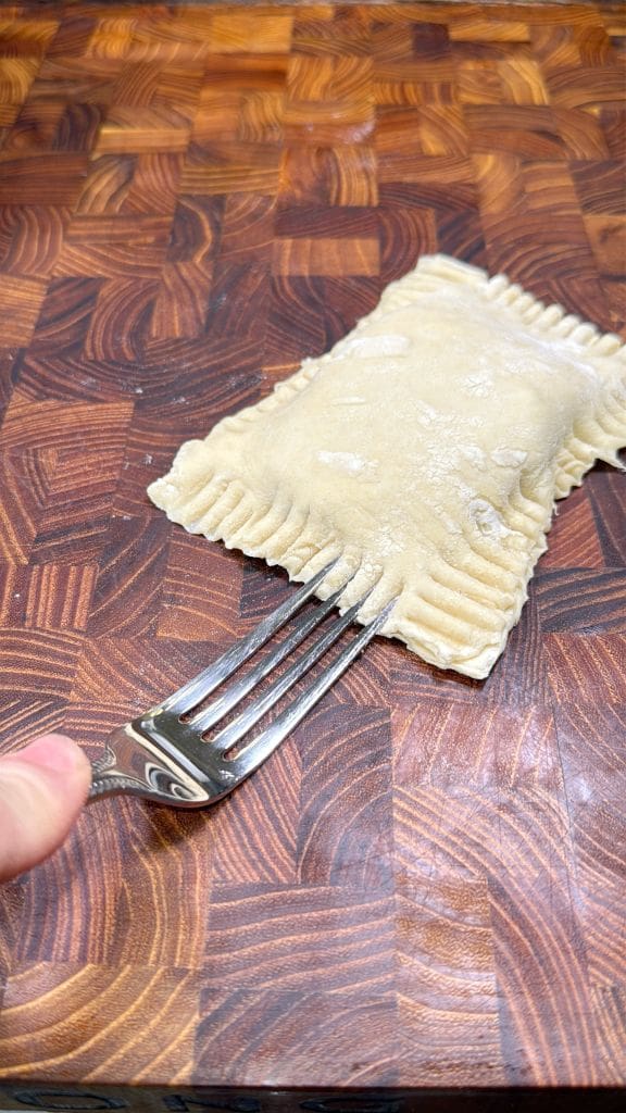 A hand holds a fork, pressing down on the edges of a rectangular piece of dough on a wooden cutting board. The dough has a crimped border, suggesting preparation of a filled pastry or ravioli.