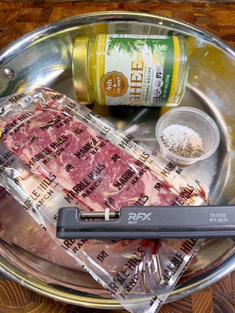 A stainless steel pan holds a vacuum-sealed package of raw meat labeled Marble Hills Ranch, a jar of Fourth & Heart ghee with a vanilla flavor, a small container of salt and pepper, and a meat thermometer.