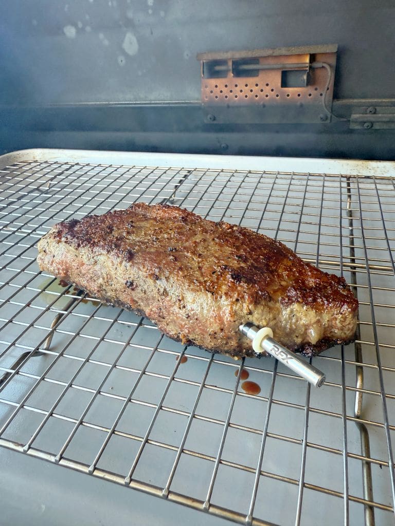 A cooked steak resting on a wire rack inside a grill, with a thermometer inserted into the side. The meat is browned with a slightly charred crust, and the grill interior is visible in the background.