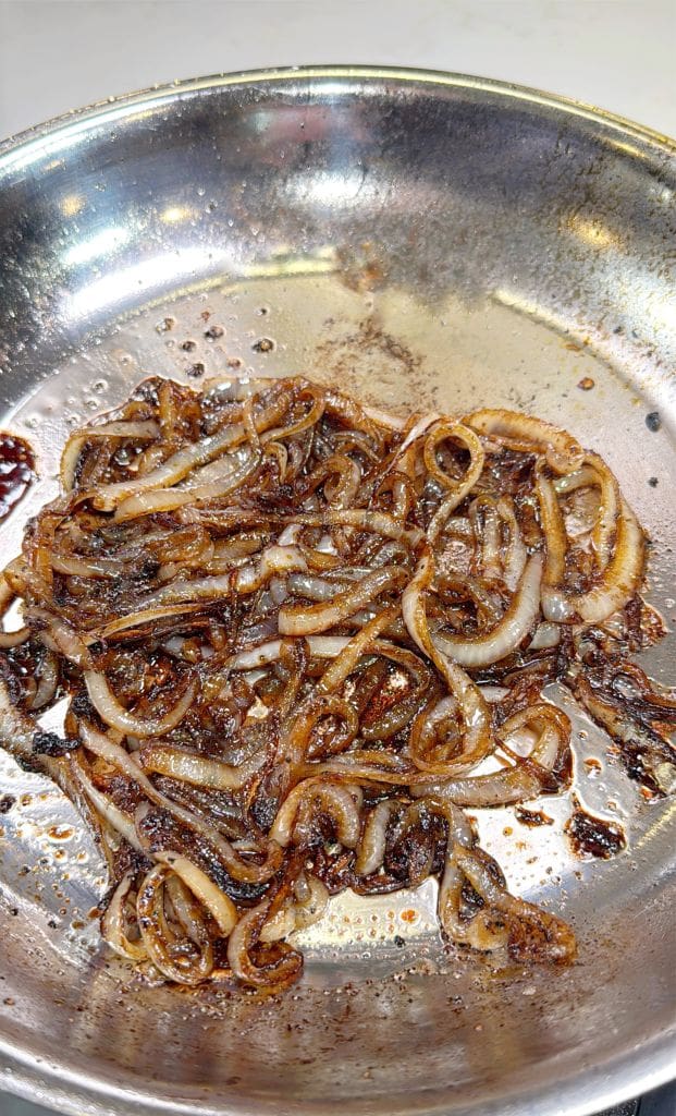 Sautéed onions in a stainless steel pan, glistening with oil and browned to a caramelized texture.