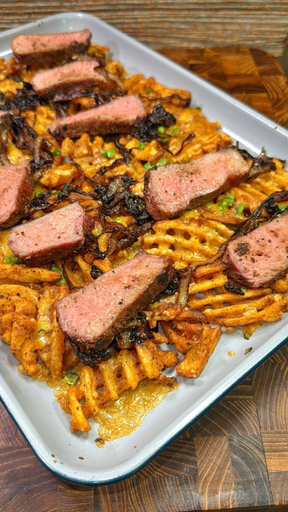 A tray of waffle fries topped with sliced steak, garnished with caramelized onions and chopped parsley. The steak is cooked to medium doneness, and the fries are golden and crispy. The background shows a wooden surface.