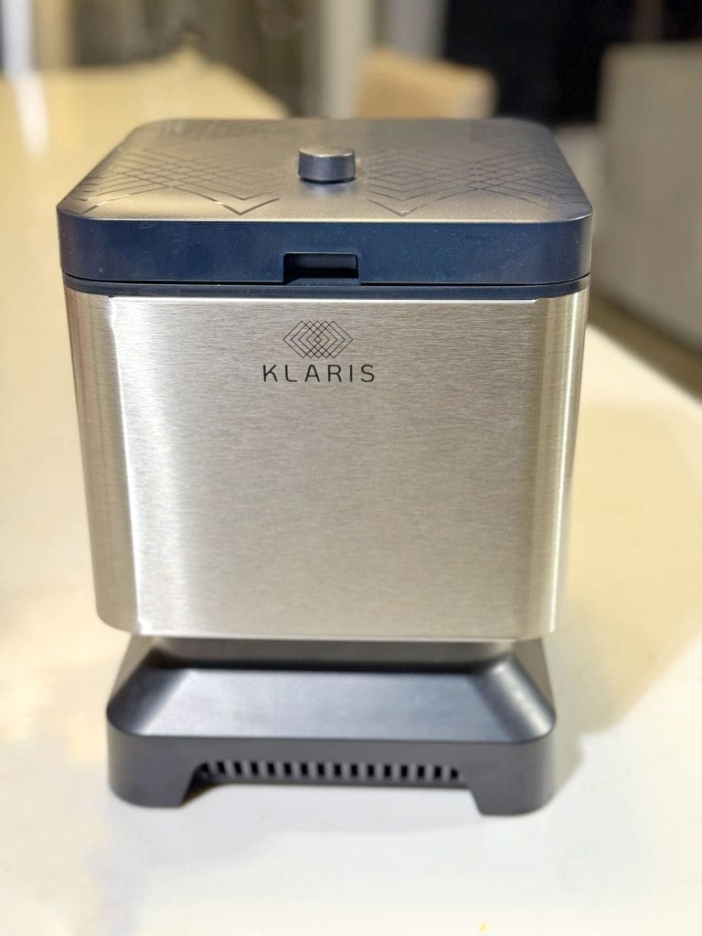 Stainless steel kitchen appliance with a dark blue lid, labeled Klaris, on a white countertop. Features a geometric design on the top and a vented base.