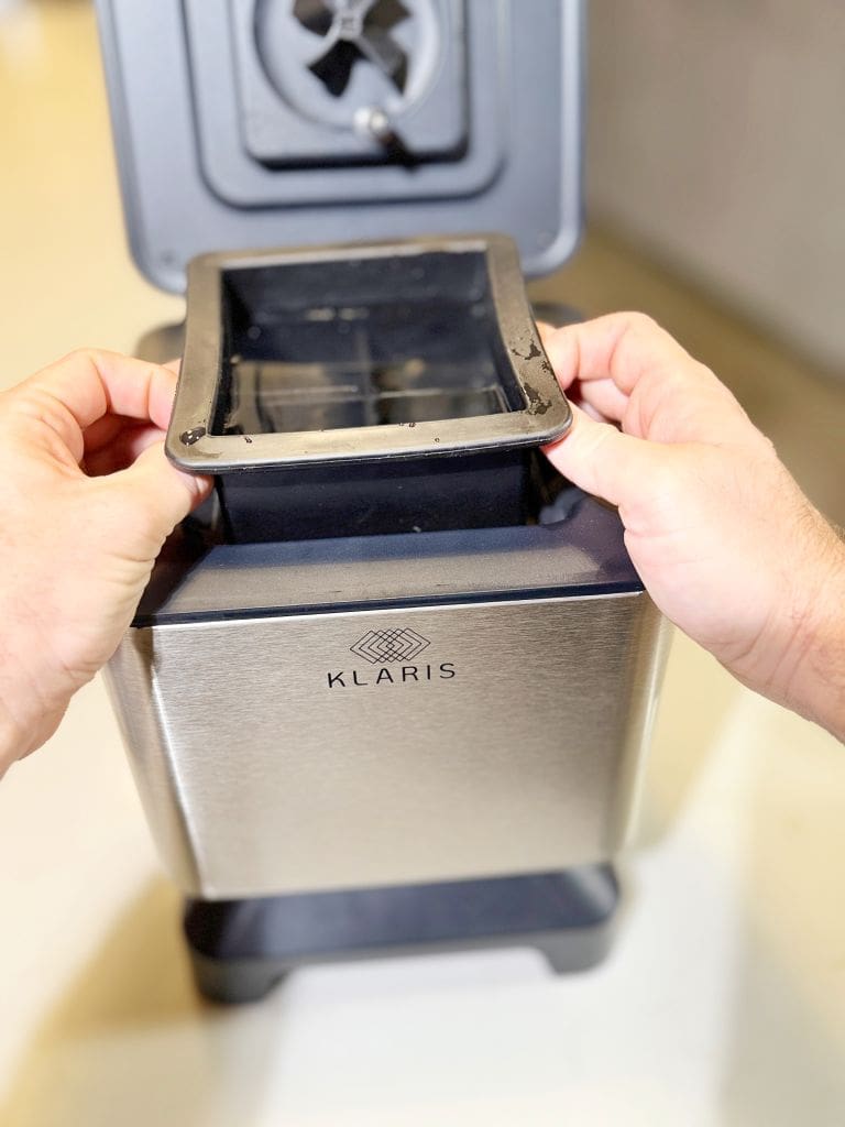A person is holding the lid of a stainless steel appliance with the brand name Klaris visible on the front. The appliance, possibly a juicer or blender, has an open top, and the background is a blurred indoor setting.