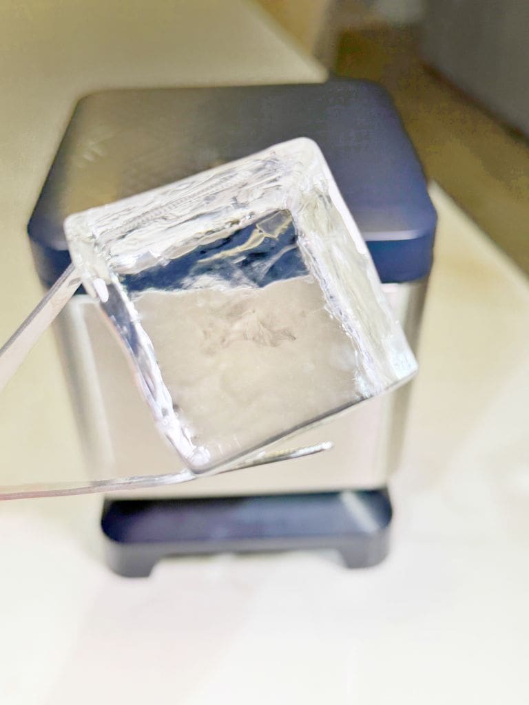 Close-up of a perfectly clear ice cube held by tweezers. The ice cube is in focus, showcasing its transparency, while the background appears slightly blurred, featuring what seems to be a silver and black appliance.