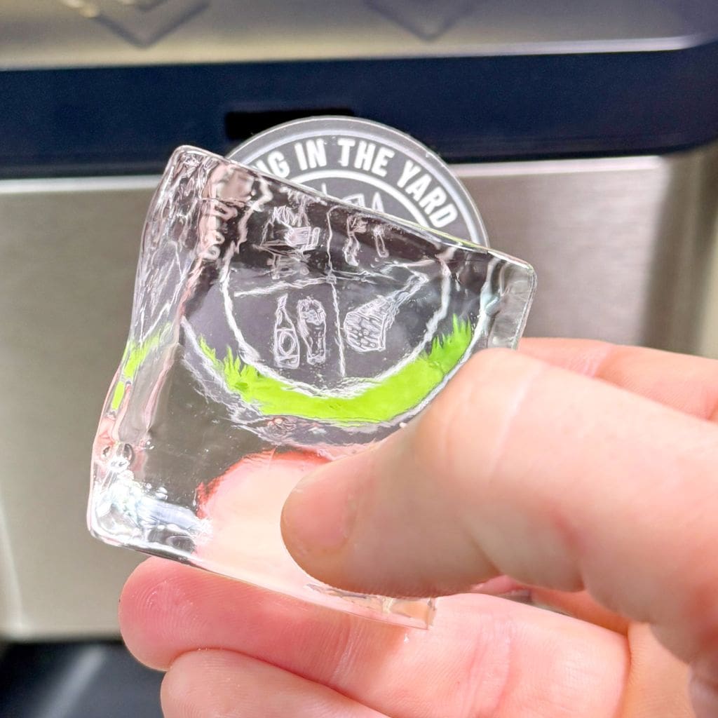 A hand holding a clear ice cube against a backdrop with a logo. Inside the ice cube is an intricate, artistically carved design featuring a bottle and other abstract shapes with a thin, bright green streak.