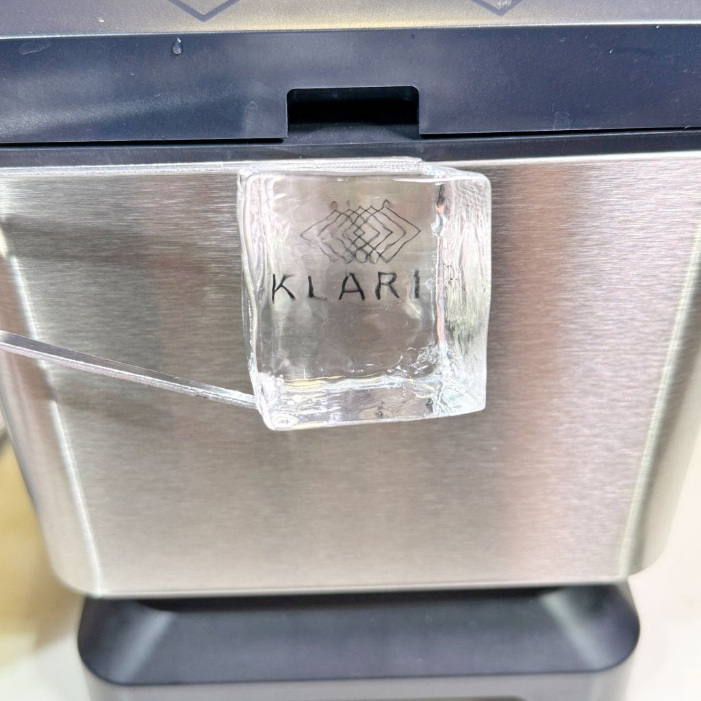 Close-up of a clear ice cube with the word Klar embossed on it. The ice cube is held by a pair of tongs in front of a stainless steel appliance, possibly an ice maker or refrigerator.