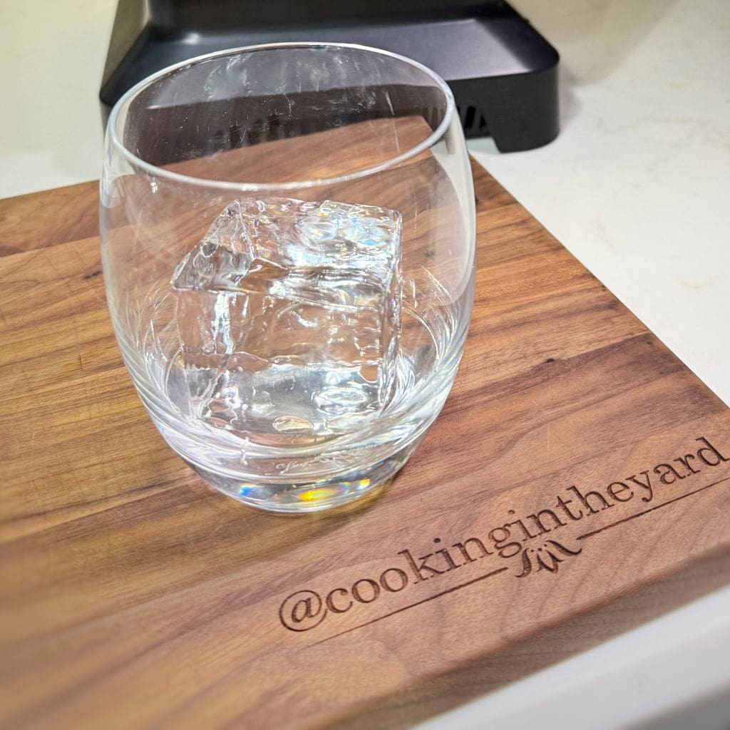 A clear glass with a large, square ice cube inside sits on a wooden cutting board. The board has the engraved text @cookingintheyard and its placed on a light-colored countertop.