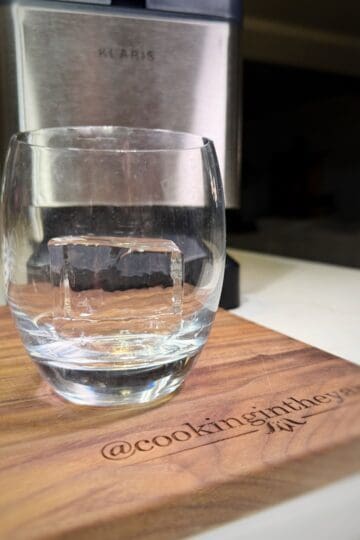 A clear glass with a large square ice cube inside rests on a wooden board. The board is engraved with @cookingintheyard. A metallic appliance is blurred in the background.