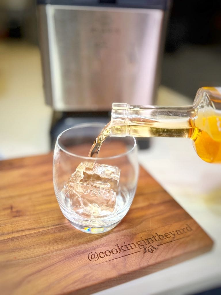 A glass with a large ice cube on a wooden board is being filled with a golden liquid poured from a bottle. The board has the text @cookingintheyard engraved on it. A metallic appliance is blurred in the background.