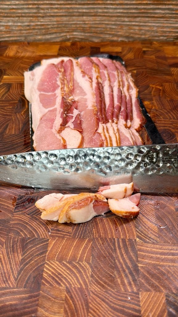 A close-up of a large knife slicing through several layers of raw bacon on a wooden cutting board. The bacon is marbled with fat, and a few pieces are cut off in front of the blade. The board has a checkered pattern.