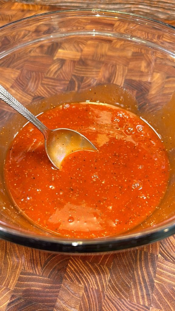 A glass bowl filled with a vibrant red sauce sits on a wooden surface. The sauce is speckled with black pepper and a metal spoon rests inside the bowl, partially submerged.