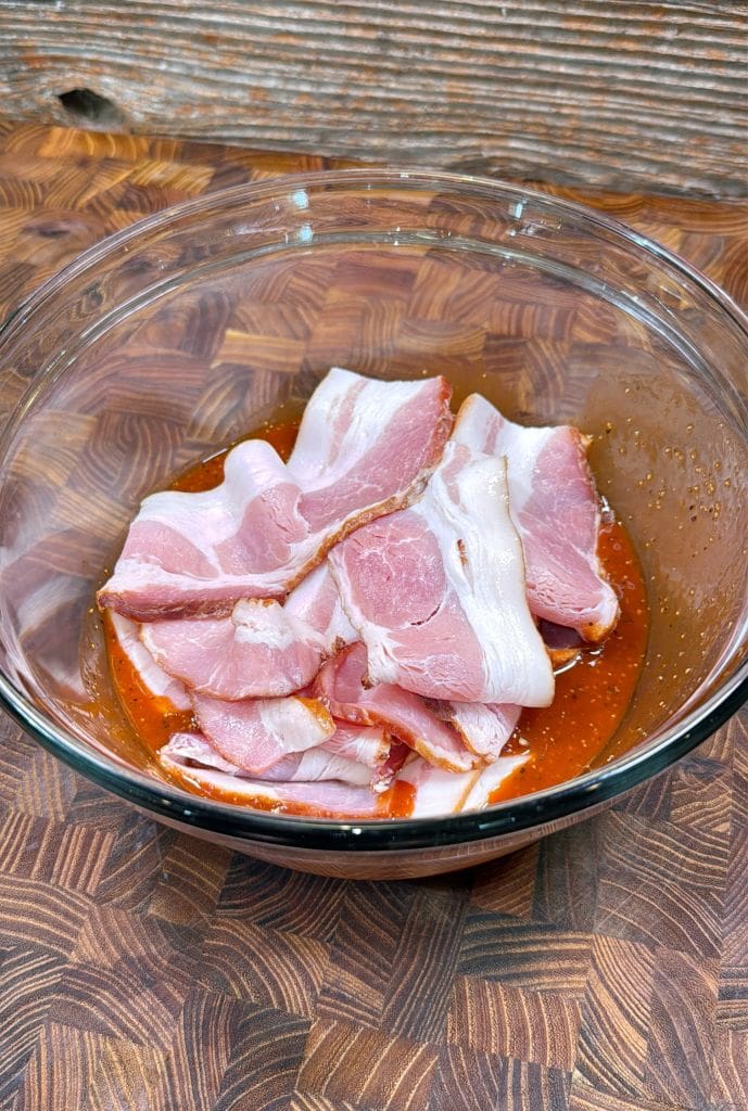 Slices of raw bacon marinating in a glass bowl with a reddish sauce. The bowl rests on a wooden surface with grain patterns.