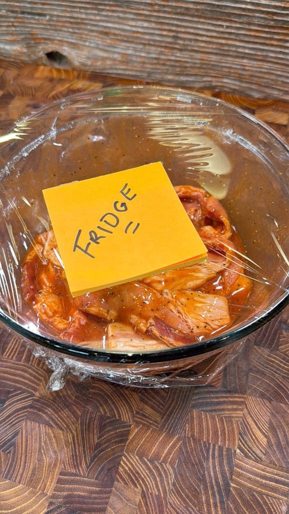 A glass bowl of food covered with plastic wrap, placed on a wooden surface. On top, a yellow sticky note labeled FRIDGE is attached to the plastic.