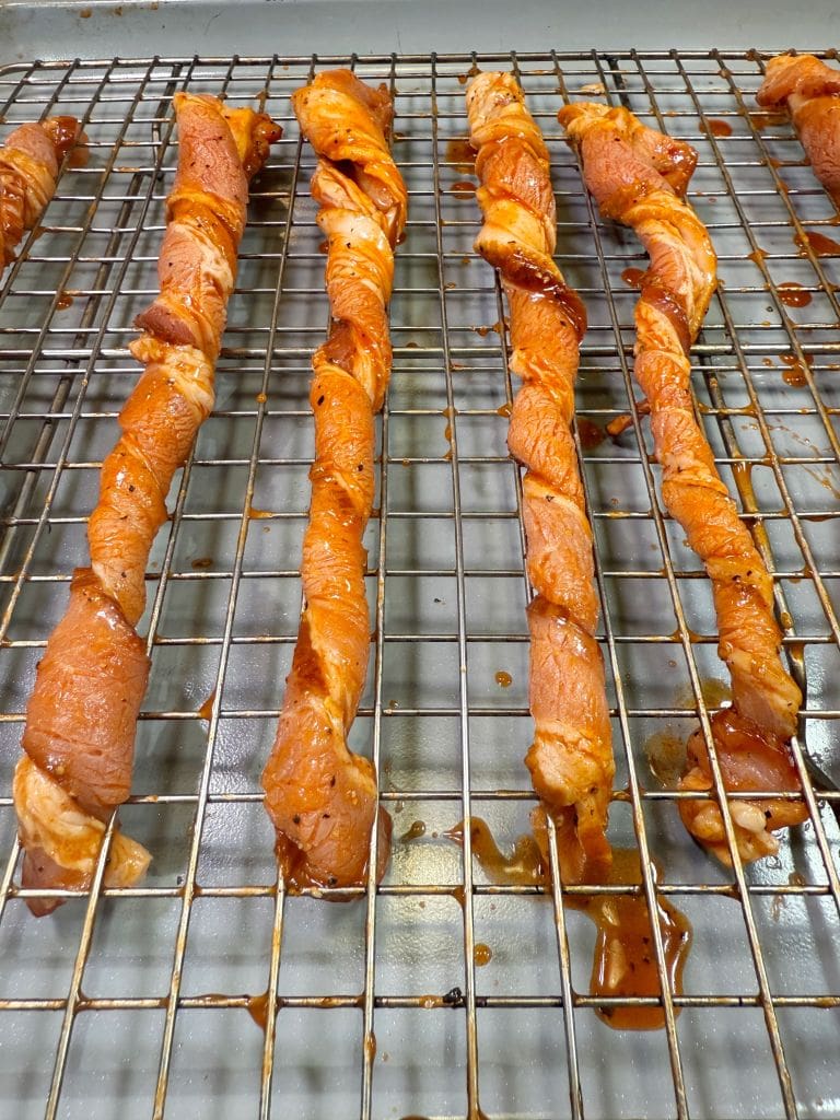 Raw, seasoned chicken skewers twisted on skewers are placed on a metal rack ready for cooking. The marinade is visibly coating the meat, giving it a shiny appearance. The rack is positioned on a baking tray.