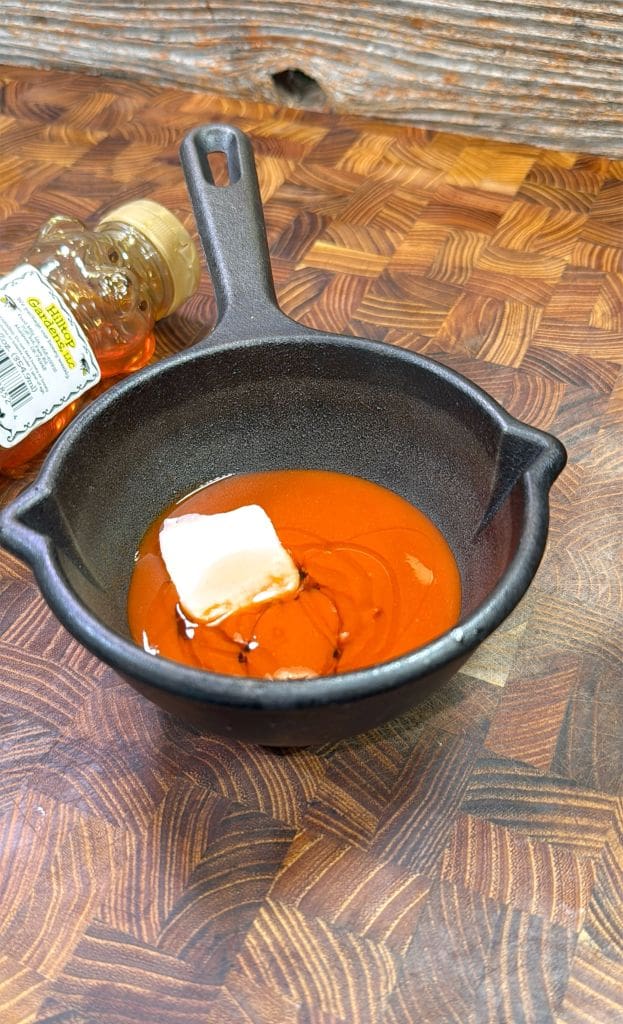 A black cast iron pan with a small block of butter and honey on a rich orange sauce, sitting on a wooden surface. A plastic honey bottle lies beside the pan.