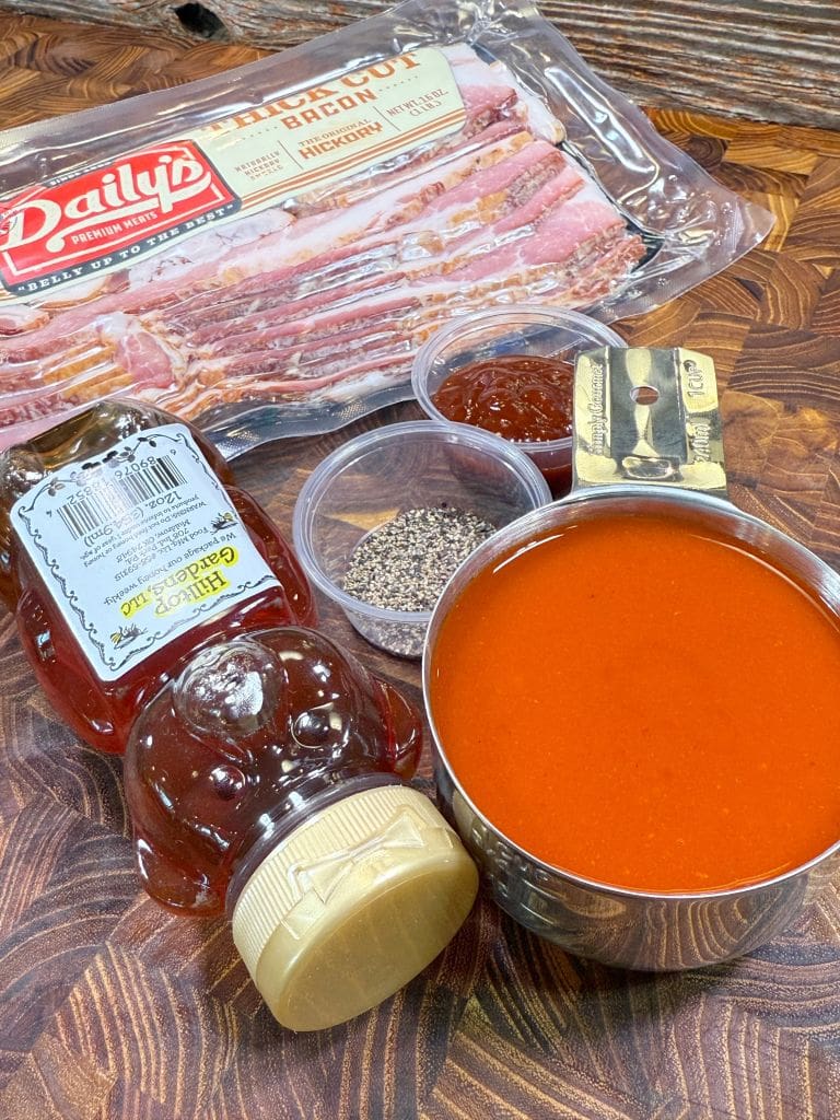 An arrangement of cooking ingredients includes a package of sliced bacon, a bottle of honey, a measuring cup filled with sauce, a small container of black pepper, and another with sauce. A razor paint scraper is also present on the wooden surface.  Ingredients are to make honey buffalo bacon twists