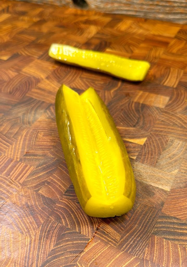 A sliced dill pickle on a wooden cutting board. The foreground features half of the pickle with seeds visible and the background shows the other half. The board has a wavy, patterned surface.