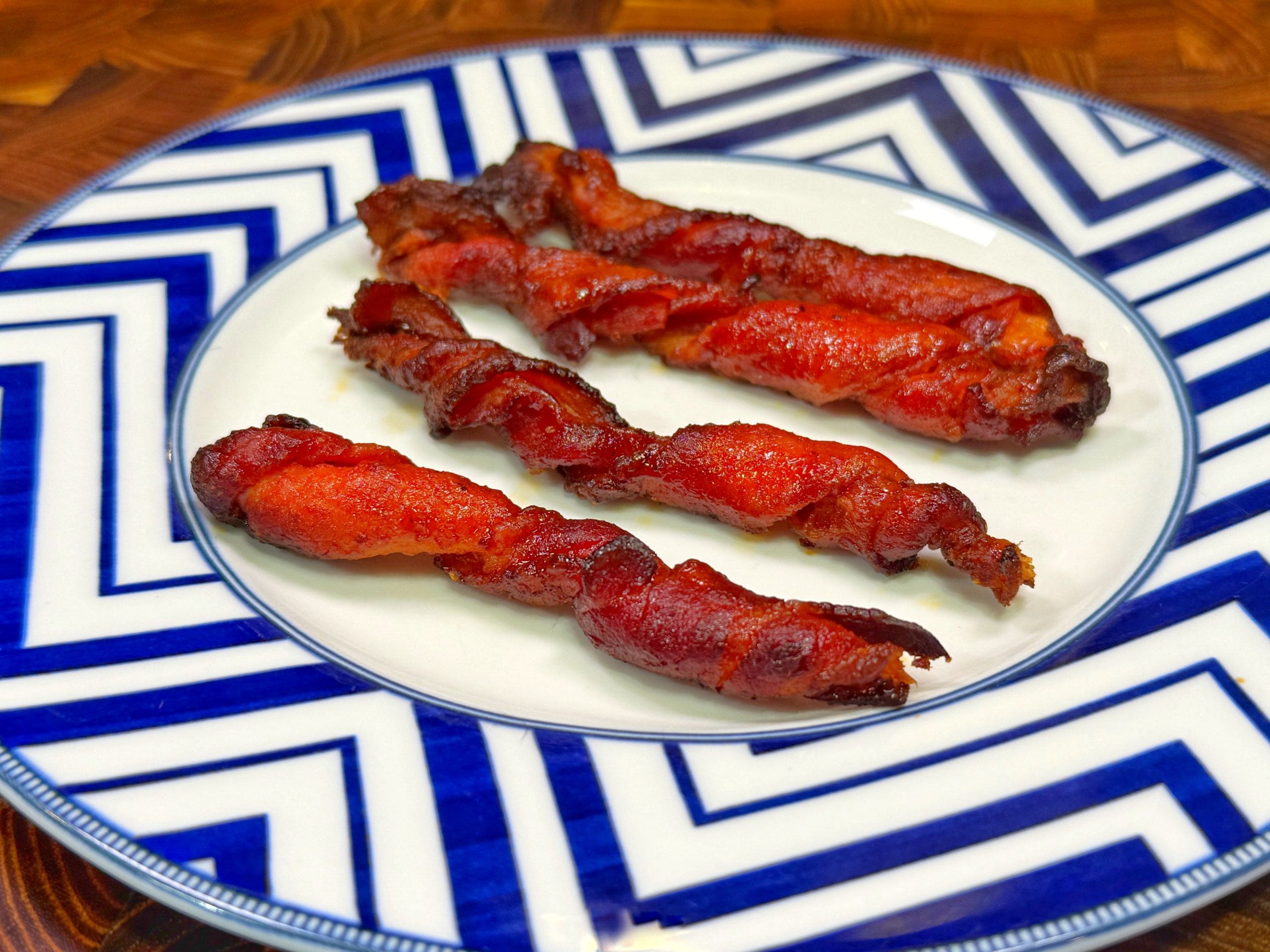 Three strips of bacon, with two twisted and one flat, on a white plate featuring blue zigzag patterns. The background shows a wooden surface.