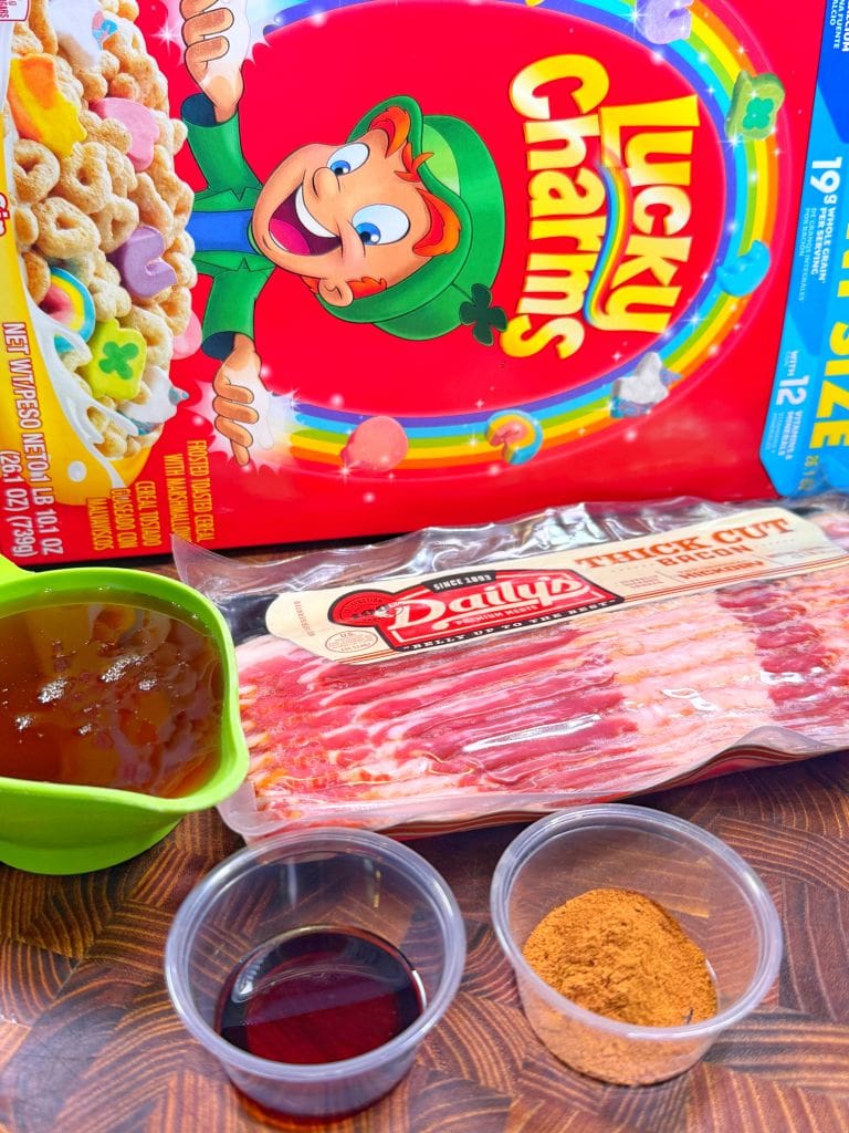 A box of Lucky Charms cereal, a package of bacon, a small cup of cinnamon, a green bowl of syrup, and a small cup of dark liquid are placed together on a wooden surface. Ingredients to make lucky charms candied bacon