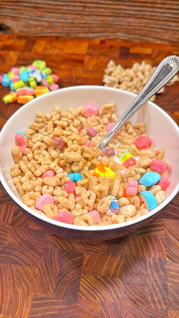 A bowl of cereal with colorful marshmallows and milk, sitting on a wooden surface. A silver spoon is in the bowl. In the background are scattered piles of multi-colored marshmallows and cereal pieces.