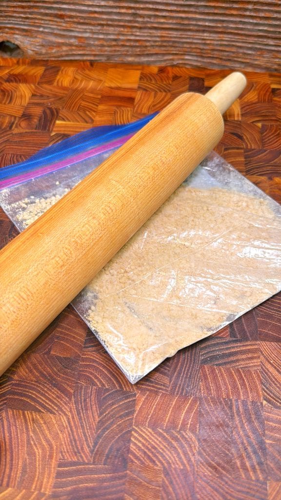 A wooden rolling pin is placed on top of a clear plastic bag filled with crushed graham crackers. The setup is on a textured wooden surface.