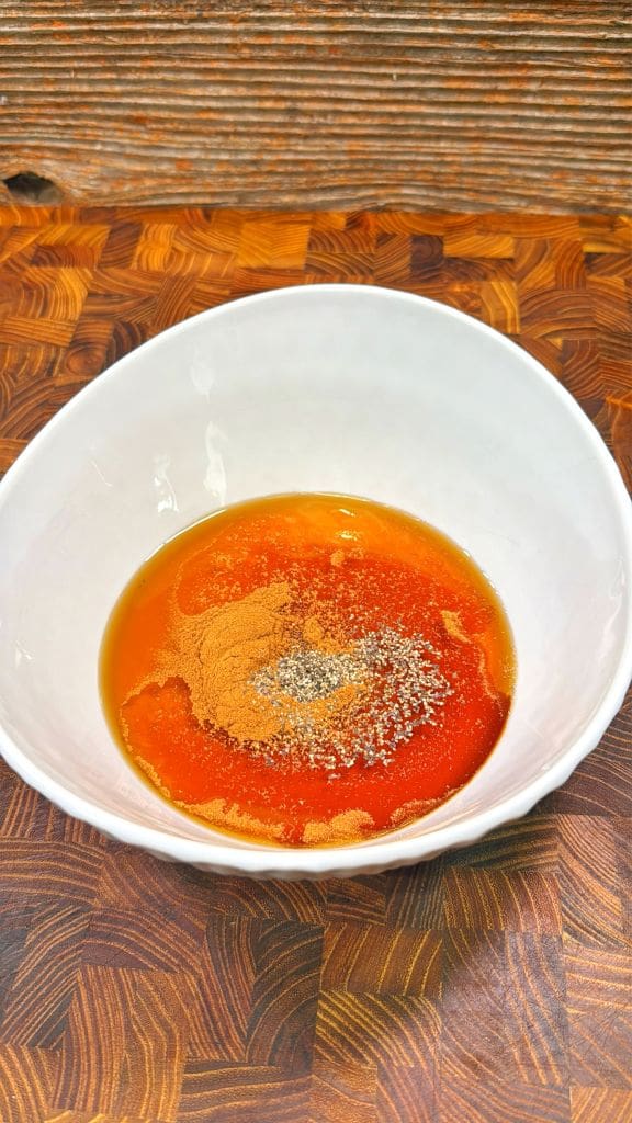 A white bowl contains a mixture of liquids and spices, including olive oil, soy sauce, paprika, and black pepper, on a wooden surface.
