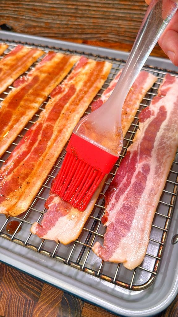 Strips of bacon are laid out on a metal rack. A hand uses a red silicone brush to apply a brown sauce onto the bacon. The rack is placed on a baking sheet, ready for cooking.