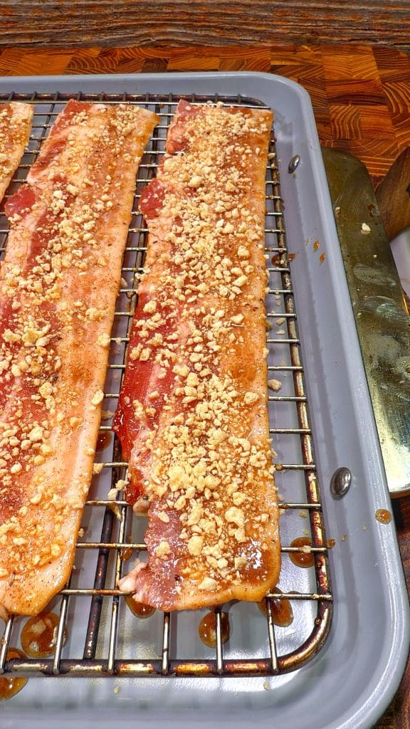 Slices of bacon covered in seasoning and crushed nuts are placed on a wire rack over a baking tray, ready to be cooked.