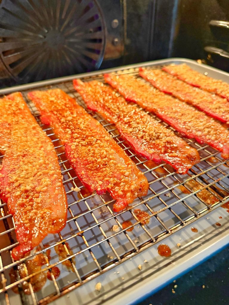 Strips of bacon covered in a reddish seasoning with sesame seeds lie on a wire rack, set over a baking tray in an oven. The bacon is evenly spaced and appears to be in the process of baking.