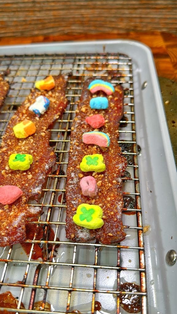 Two strips of bacon on a wire rack, topped with colorful marshmallow cereal pieces in various shapes, including stars and clovers. The bacon appears caramelized on a metal baking sheet.
