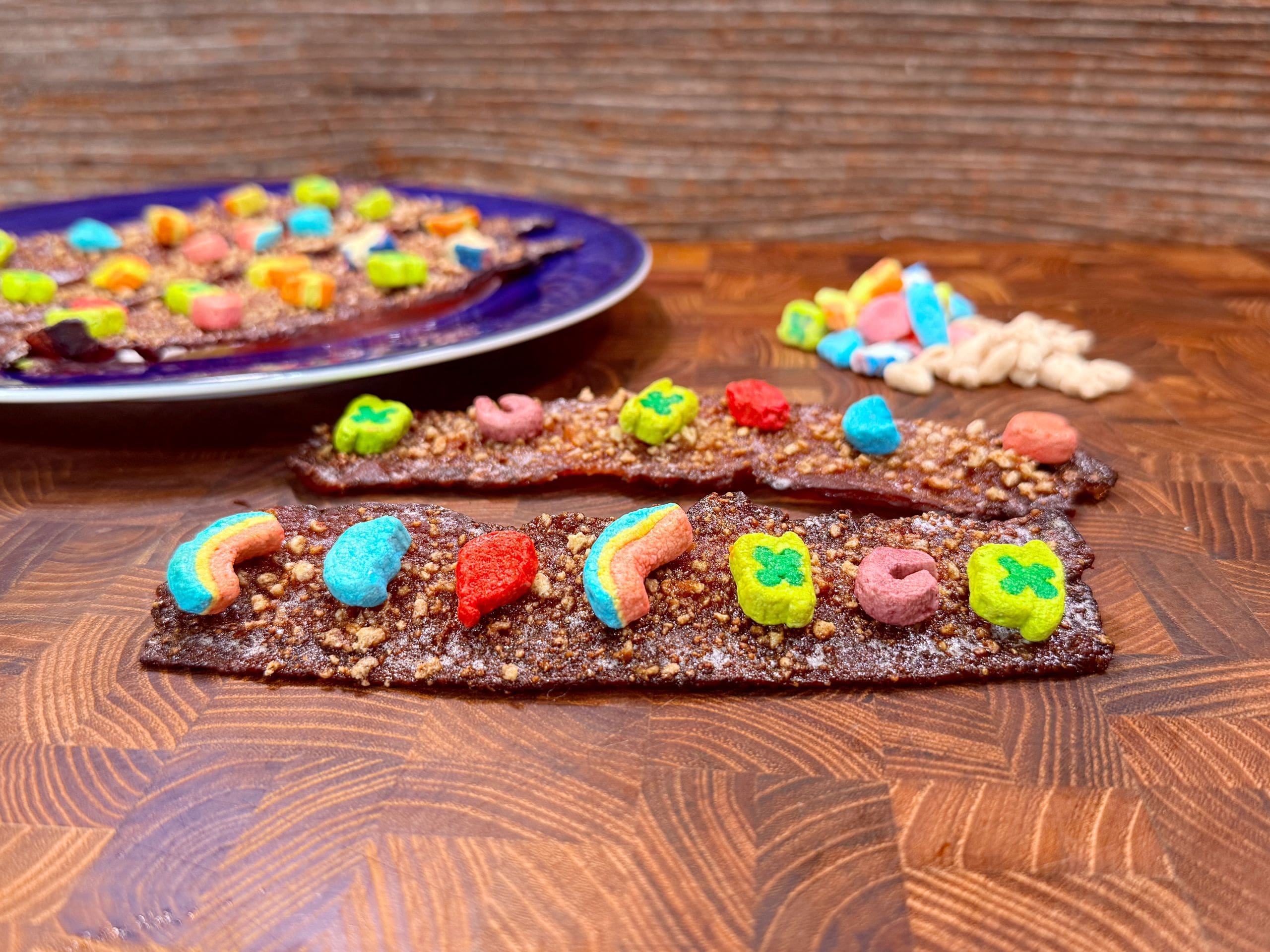 Two chocolate-covered strips topped with colorful cereal pieces are shown on a wooden surface. A purple plate with more strips is in the background, alongside a few cereal pieces scattered around.
