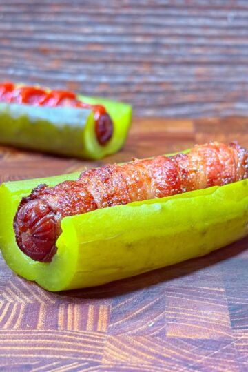 A grilled sausage wrapped in bacon is placed inside a halved pickle on a wooden surface. Another similar pickle and sausage combination is visible in the background.