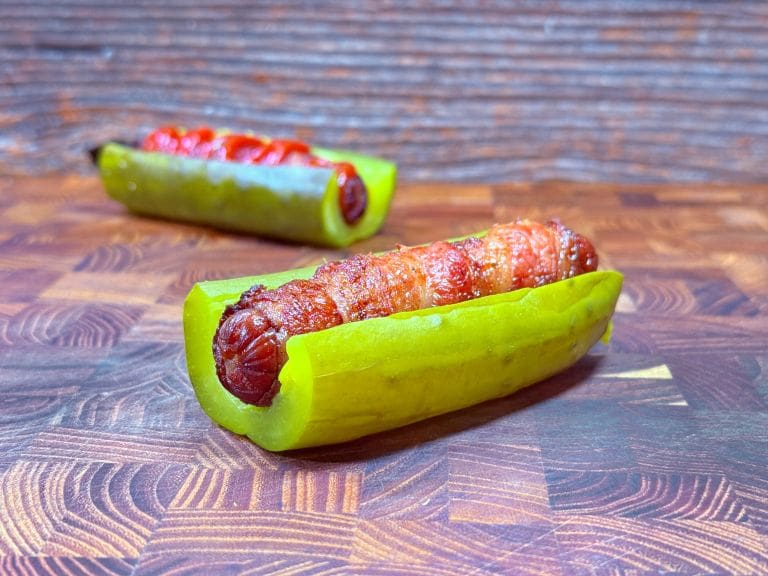 A grilled sausage wrapped in bacon is placed inside a halved pickle on a wooden surface. Another similar pickle and sausage combination is visible in the background.