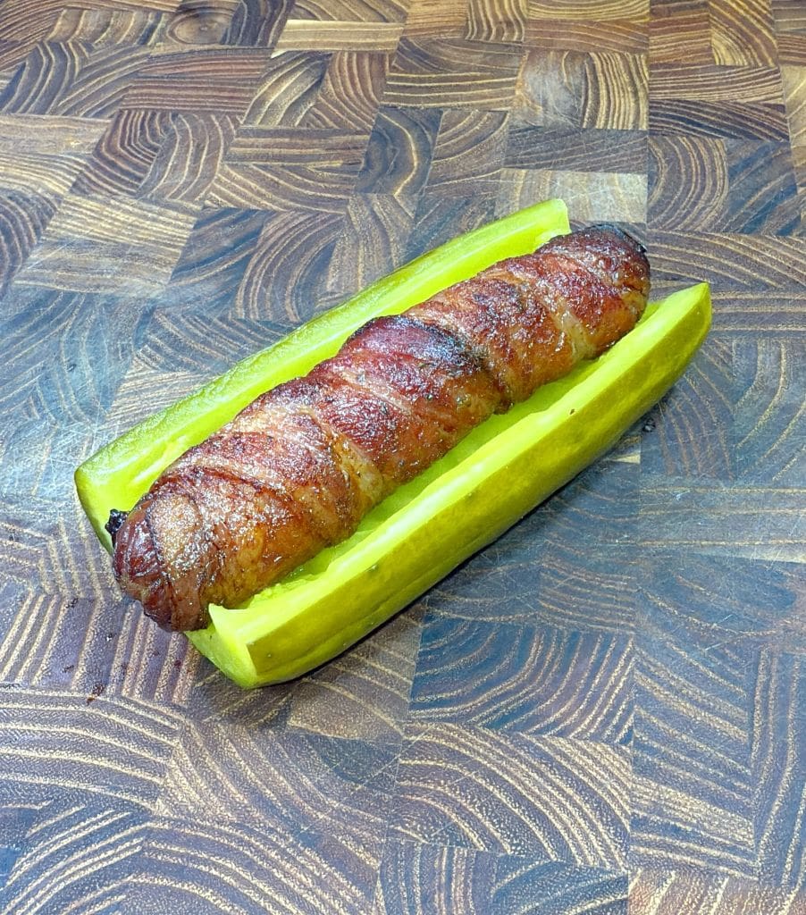 A bacon-wrapped sausage placed between two pickle halves on a wooden surface. The pickle halves mimic a bun, holding the sausage together. Picture of a bacon wrapped hot dog in a pickle hot dog bun