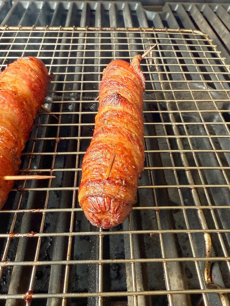 Two hot dogs wrapped in crispy bacon are placed on a grill rack. They are secured with toothpicks to keep the bacon in place. The grill marks indicate they are being cooked evenly.