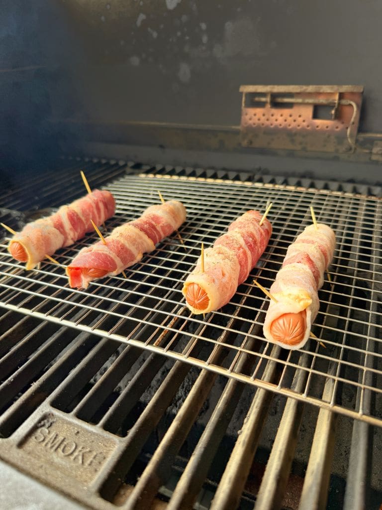 Four bacon-wrapped hot dogs secured with toothpicks are cooking on a grill grate. The grill is open, showing the smoky chamber and the sausages positioned evenly on the hot surface.