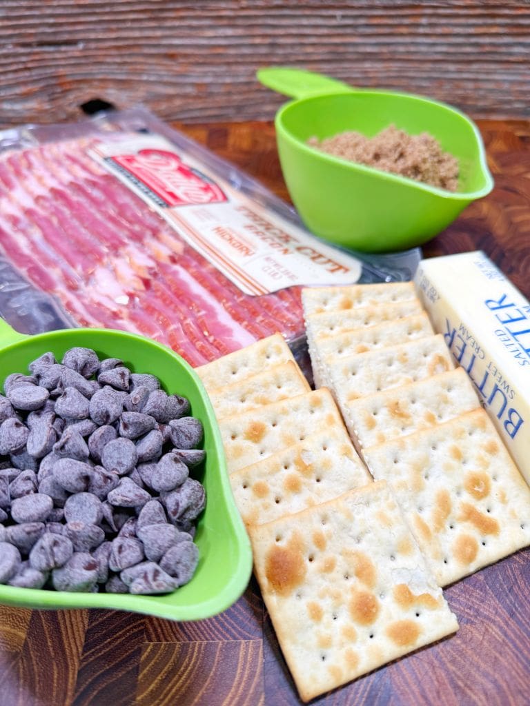 A wooden surface with ingredients including packaged bacon, a green measuring cup with brown sugar, butter, saltine crackers, and another green cup with chocolate chips.