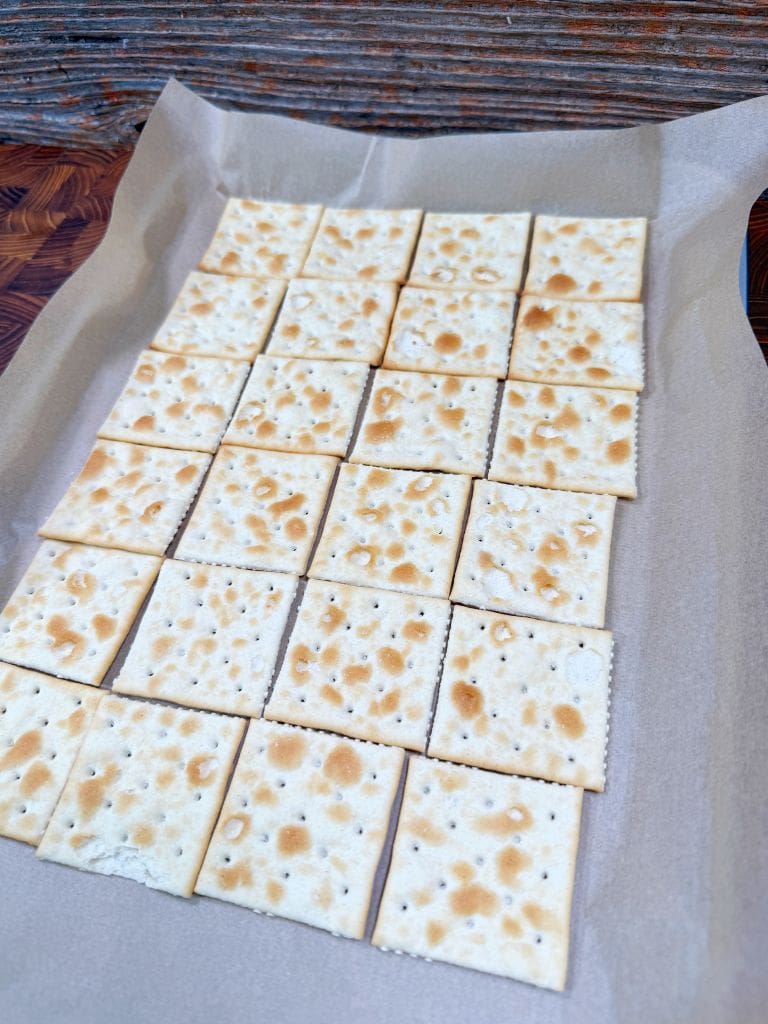 A grid of square saltine crackers arranged neatly on a sheet of parchment paper. The crackers have a light, baked texture with small holes and brown spots, set against a rustic wooden background.